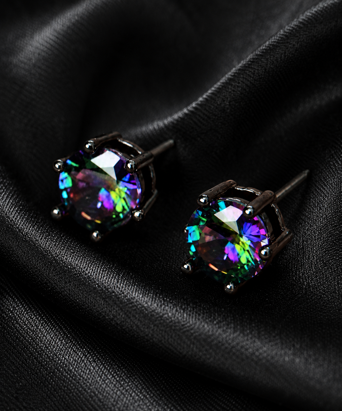The Black Doll Affair's Dark Matter ME Doll pair of round iridescent gemstone stud earrings in a dark metal setting, displayed on a draped black satin fabric background.