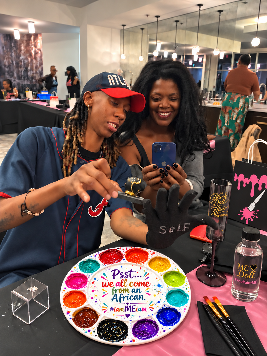 Mama Doll and a Black Doll smiling at the painted results during a ME Doll Group Experience art workshop with a colorful paint palette, painted ME Dolls and art supplies.