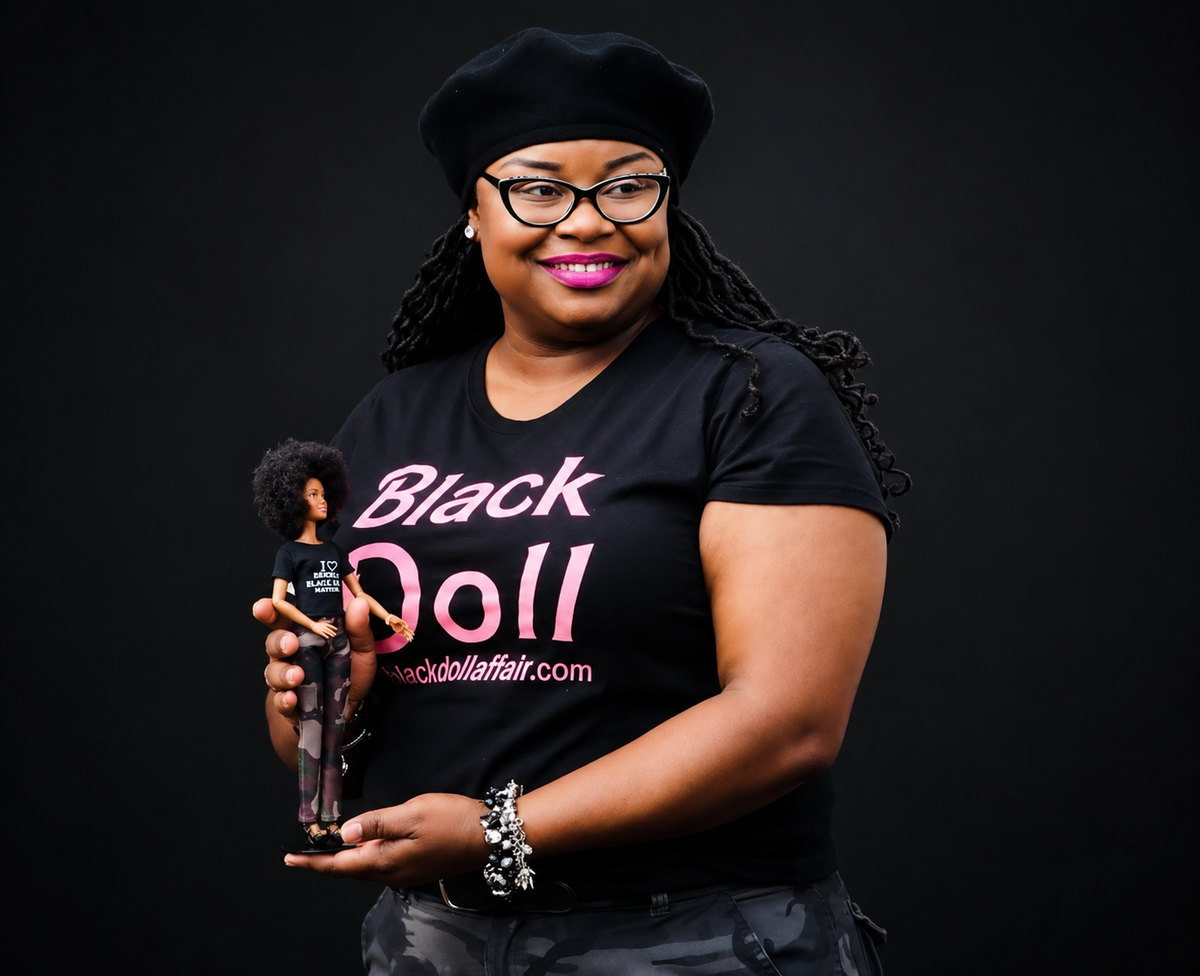 Standing against a solid Black studio background, a smiling member of the Black Doll Affair, a Black Doll, wears The Black Doll Affair Signature Members T-shirt with pink text reading 'Black Doll' and blackdollaffair.com and holds Barbie Fashionistas Doll #59 inspired by Mama Doll's advice to Mattel to expand representation and put an afro on Barbie. 