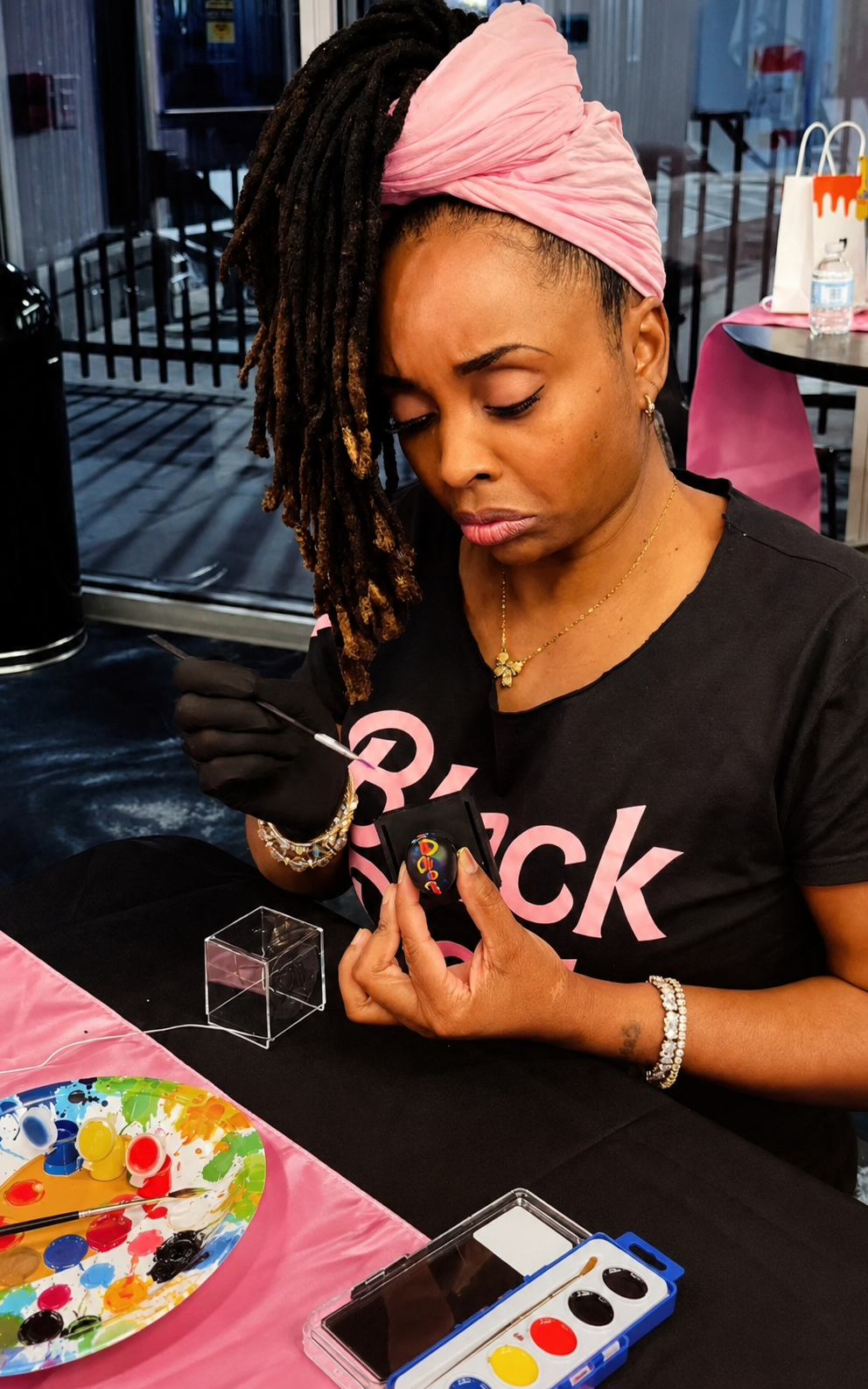 Wearing a pink head wrap and dreadlocks, Black Doll Michelle Rosser carefully hand-paints a ME doll head before winning BEST IN SHOW at The Black Doll Affair's paint & sip party. 