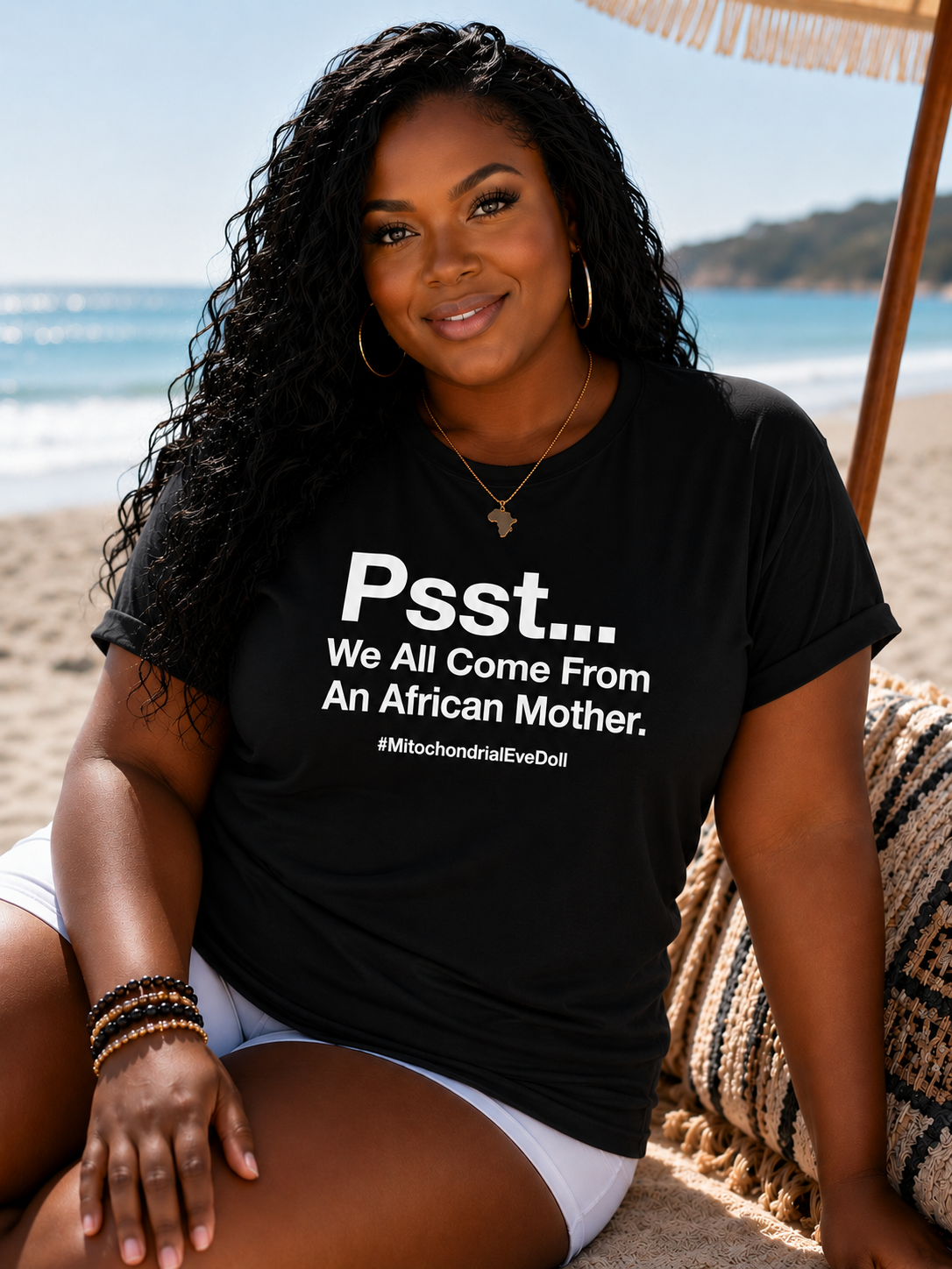 A woman sitting on a beach chair on a sandy beach wearing a black graphic t-shirt with white text that reads 'Psst... We All Come From An African Mother.