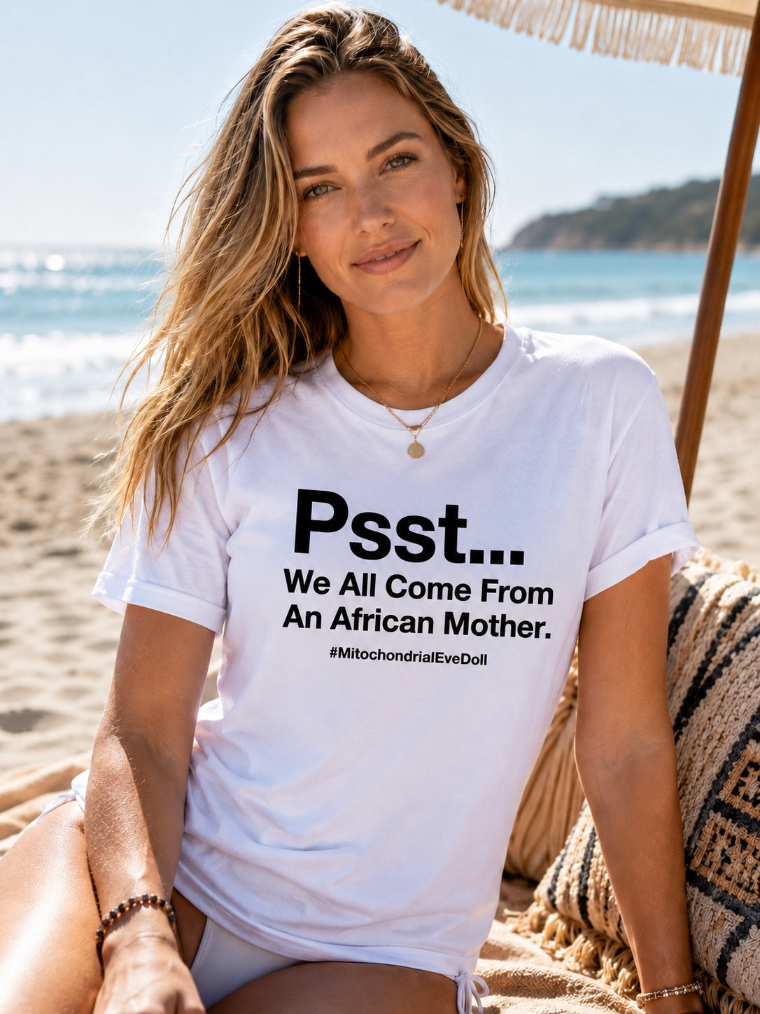 A woman sitting on a beach chair on a sandy beach wearing a white graphic t-shirt with white text that reads 'Psst... We All Come From An African Mother.
