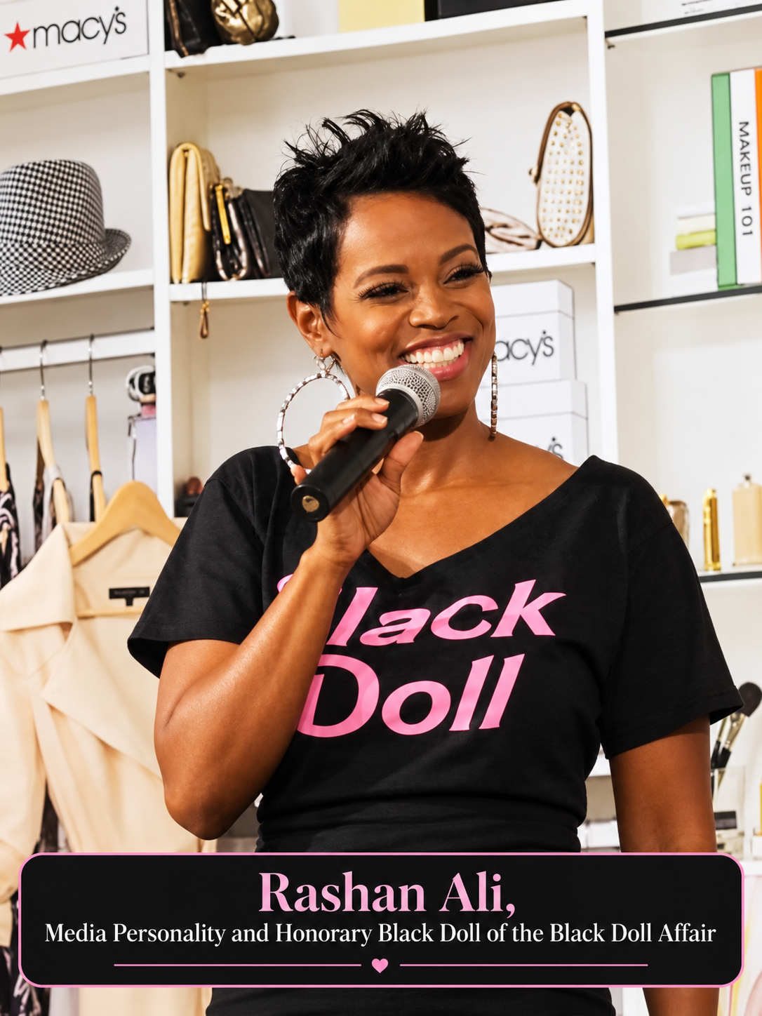 Media personality, Rashan Ali, an Honorary Black Doll of the Black Doll Affair, smiling woman with short black hair holds a microphone in front of her face while wearing a black V-neck T-shirt with 'Black Doll' printed in pink across the front. She is standing in front of white shelves displaying cosmetics and accessories, with a Macy's logo visible on the shelf.