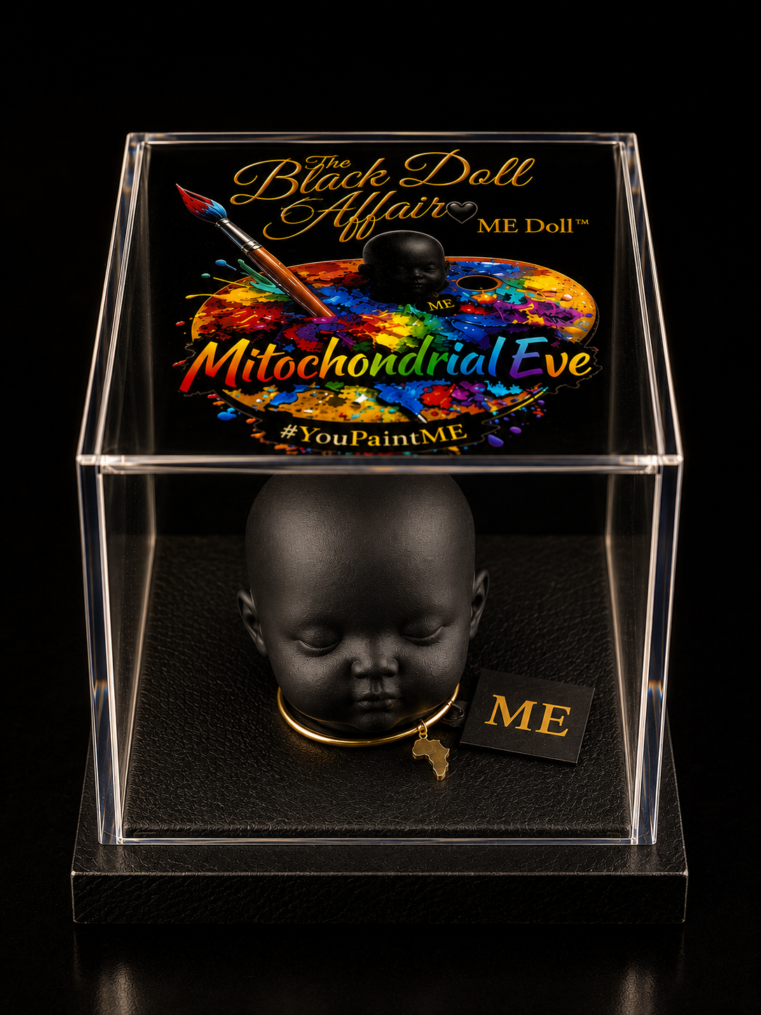 Matte-black ME Doll head wearing the signature gold necklace and ME neck tag with a gold map of Africa charm, presented inside a clear acrylic display case with a colorful lid that shows The Black Doll Affair ME Doll logo design.  
